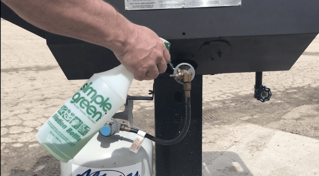 A person using a Simple Green spray bottle to check the propane connection on a grill for leaks. The propane tank and hose are visible.